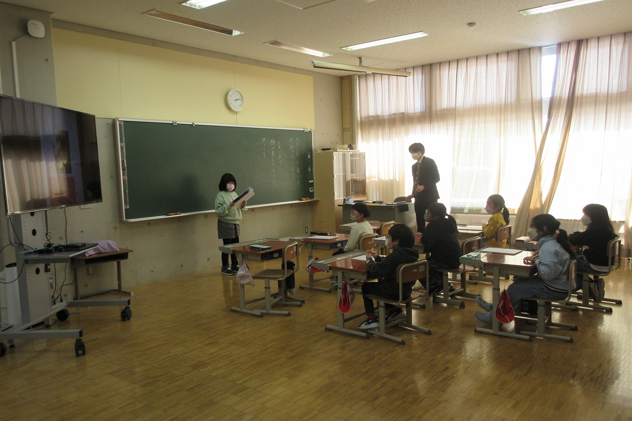 小学校の教室内にて、黒板の前で立ち、資料を持って発表を行う女子児童と、発表を椅子に座りながら聞く6人の児童の様子を写した写真