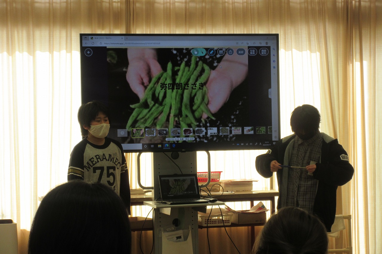小学校の教室内にて、モニターの前に立ち、モニターに映った野菜の写真を使って発表を行う、男子児童2人の様子を写した写真