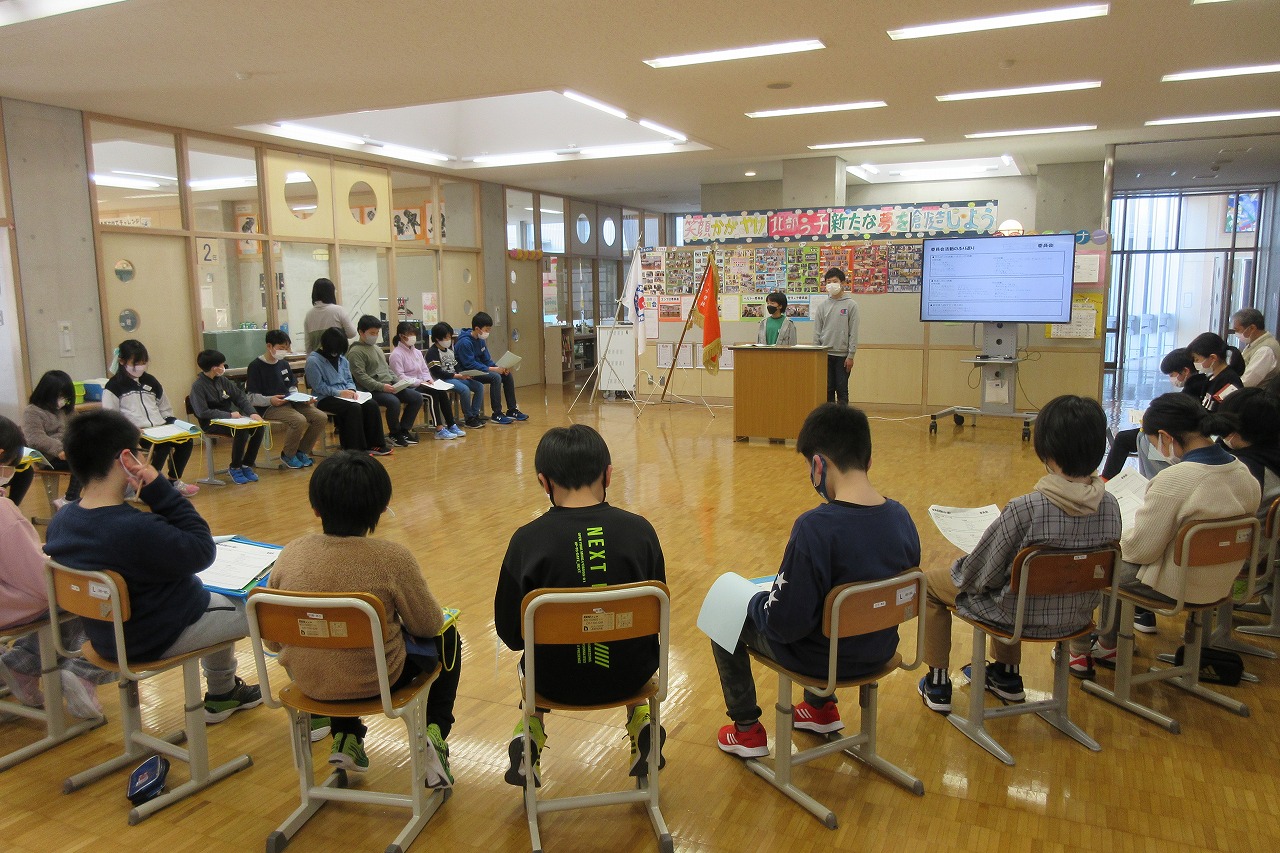 演台に立って話す2人の委員とそれを囲むように円形に並べられた椅子に座って聞く児童たちの写真