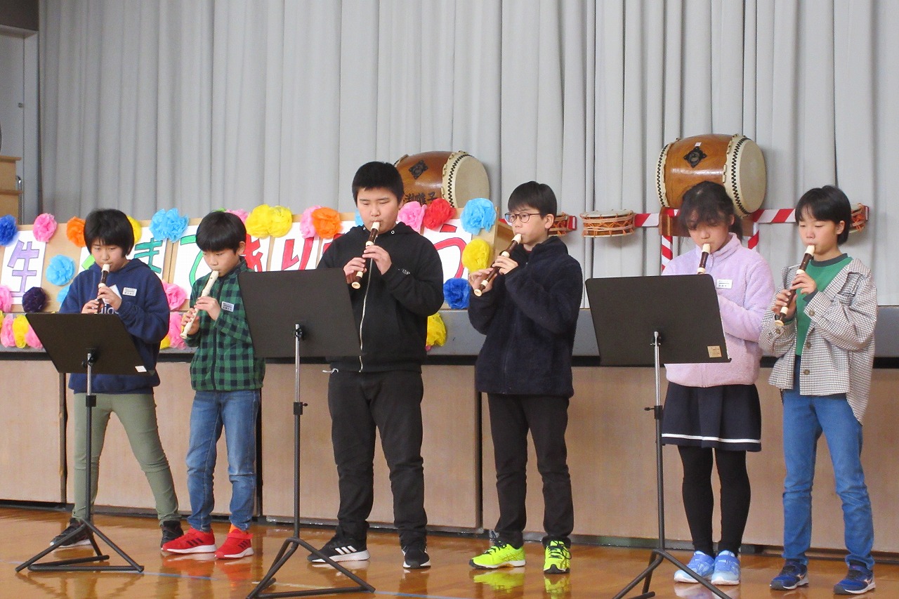 小学校の体育館にて行われた6年生を送る会にて、壇上の前でリコーダーの演奏をする、5年生の男子児童4人と女子児童2人の様子を写した写真