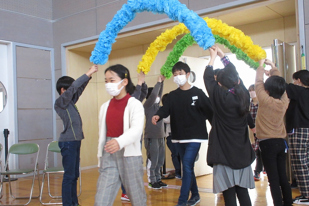 小学校の体育館にて行われた6年生を送る会にて、児童が2人一組で頭上に持ち上げて作られた、3色の花紙のアーチの下を歩いてくる、6年生の児童達の様子を写した写真