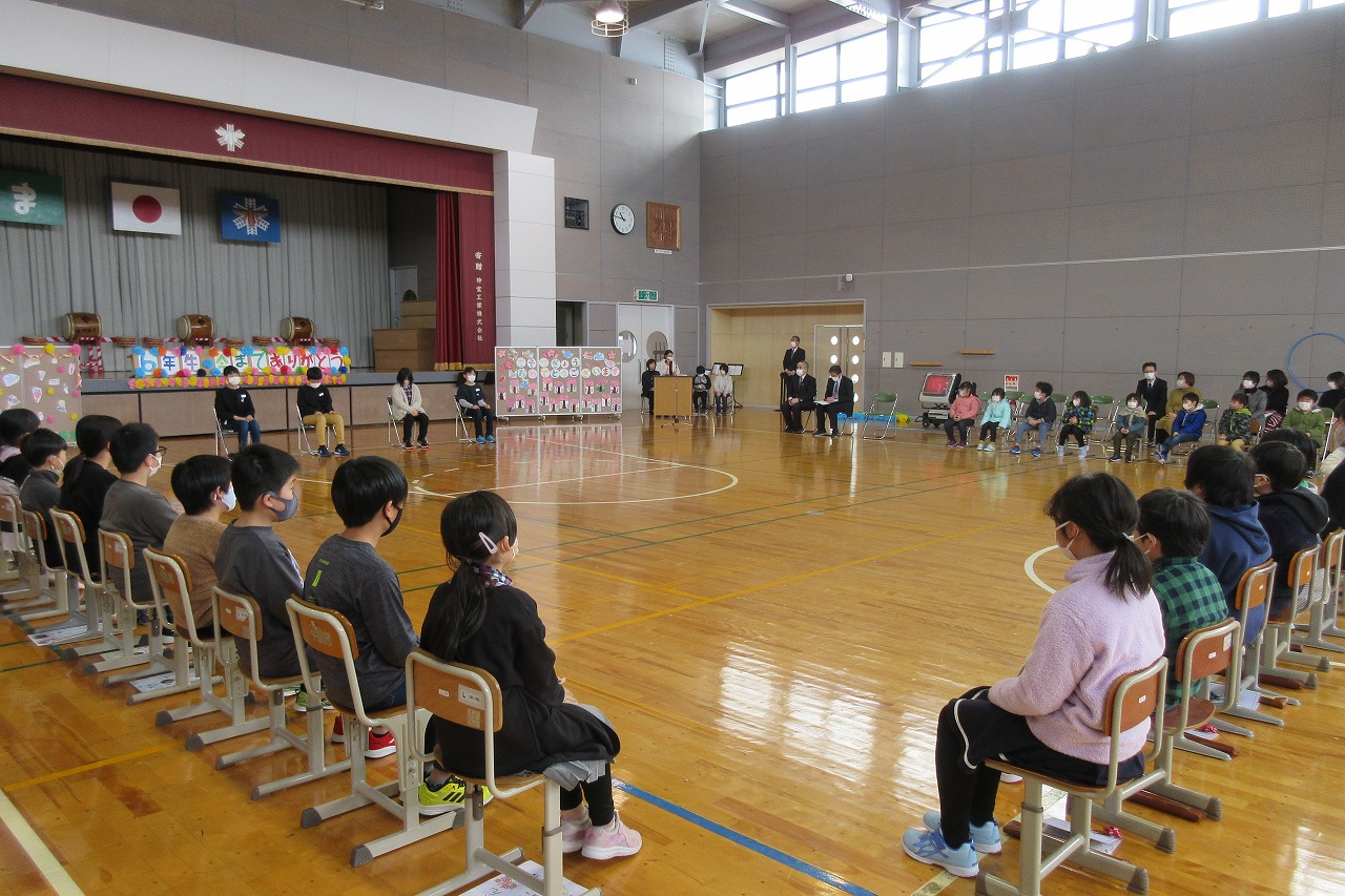 小学校の体育館にて行われた6年生を送る会にて、四角の形に並べられた学校椅子に座る、大勢の児童達の様子を写した写真