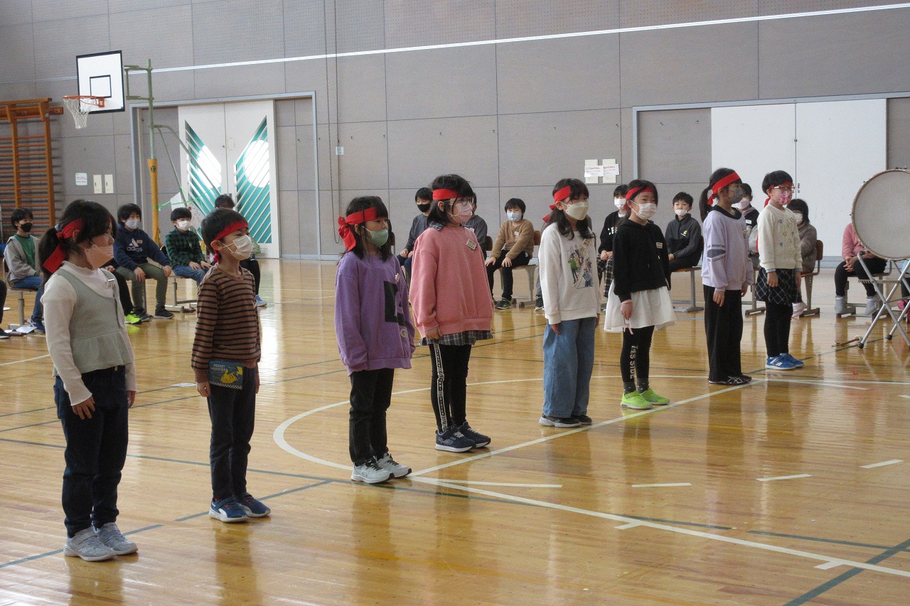 小学校の体育館にて行われた6年生を送る会にて、赤い鉢巻を巻いた8人の児童が、体育館の中央で並んで立っている様子を写した写真