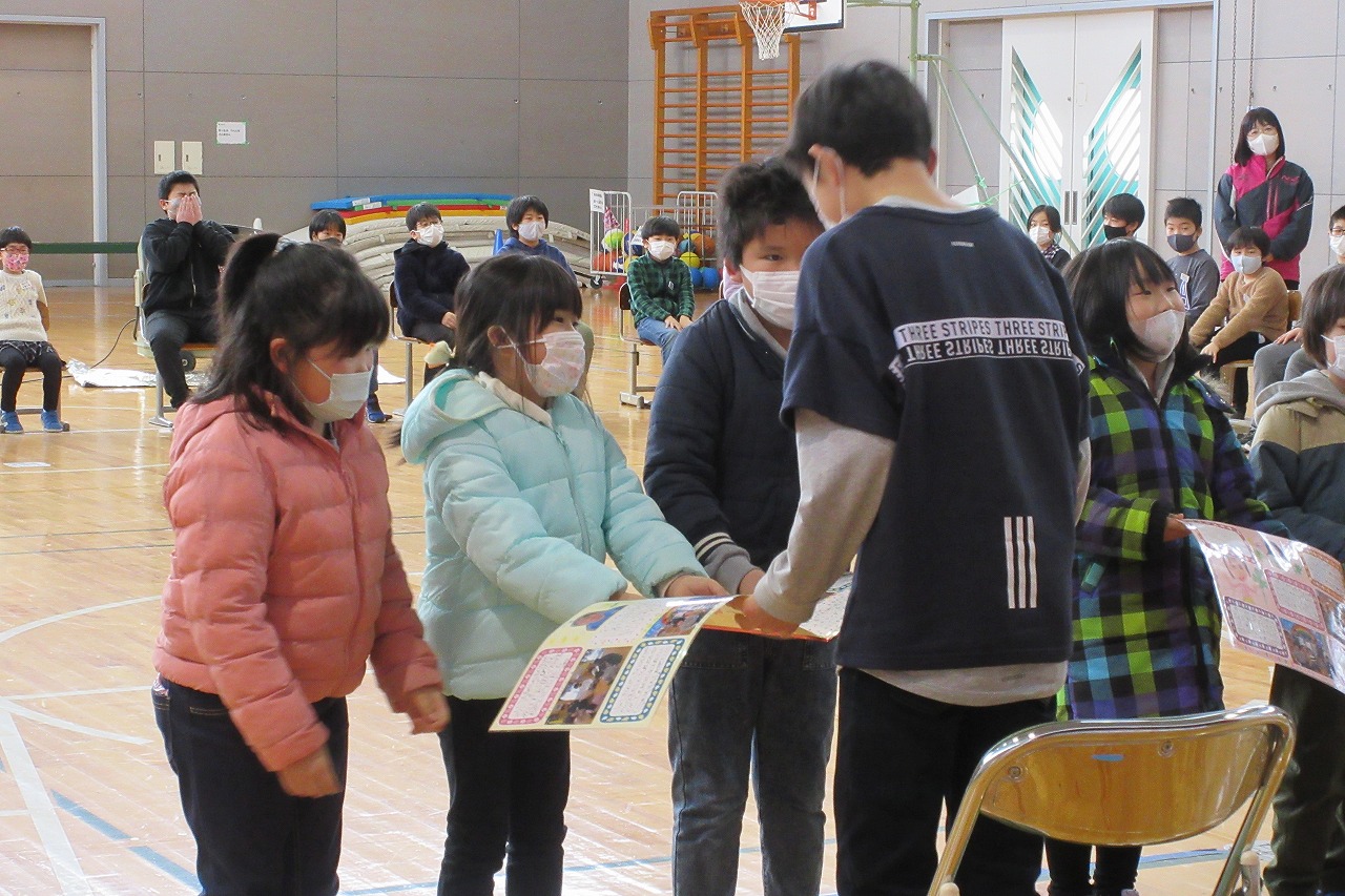 小学校の体育館にて行われた6年生を送る会にて、6年生の男子児童に記念品を贈る男子児童1人、女子児童2人の様子を写した写真