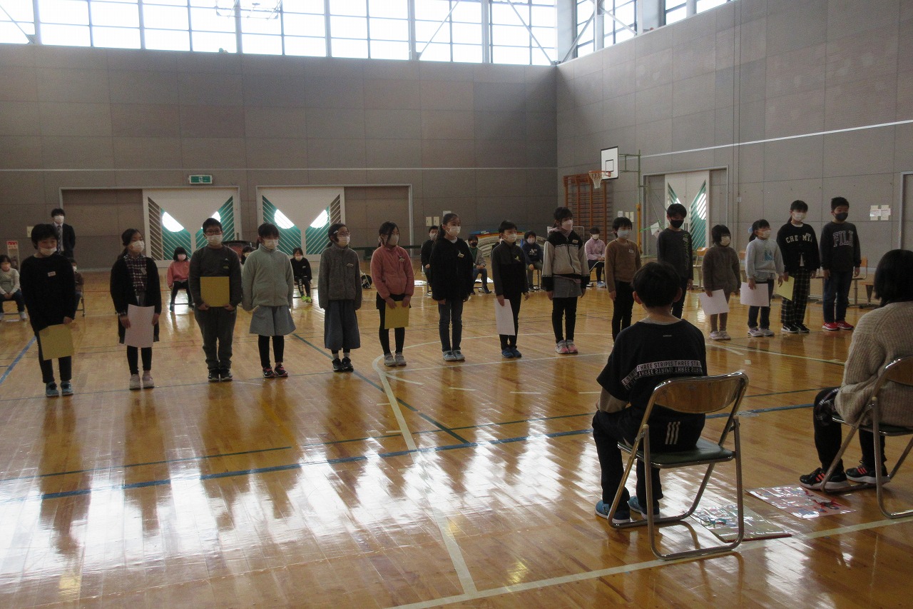 小学校の体育館にて行われた6年生を送る会にて、15人の児童が紙を持ちながら、体育館の中央で並んで立っている様子を写した写真