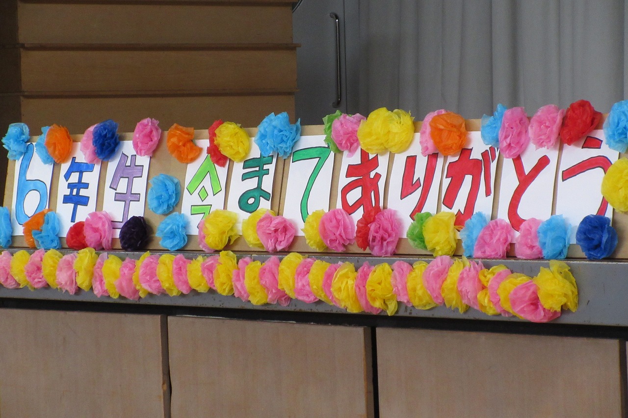小学校の体育館にて行われた6年生を送る会にて、壇上に置かれている、「6年生、今までありがとう」と7色で書かれ、その文字の周りを花紙でカラフルに彩られた作品を写した写真