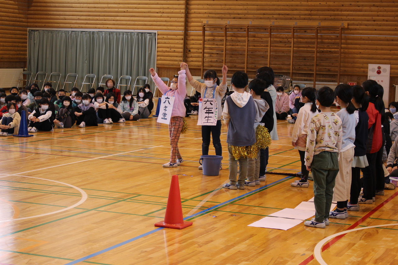「六年生」と書かれた紙を首から下げた女の子2人が万歳をし、体育座りをした子供たちがそれを眺めている写真