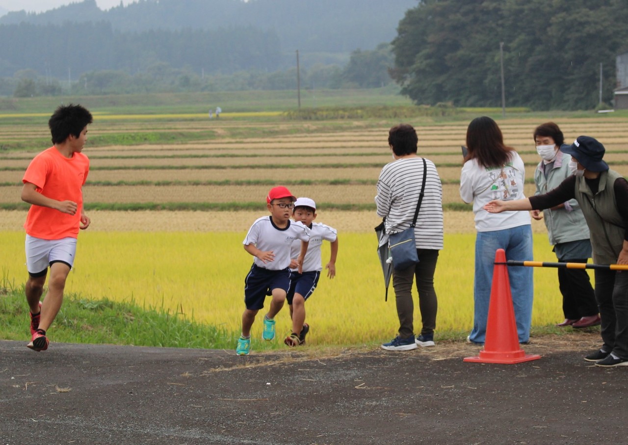 田んぼの横の道を走る体操服姿の子供に男性が並走し、それを女性たちが応援している写真