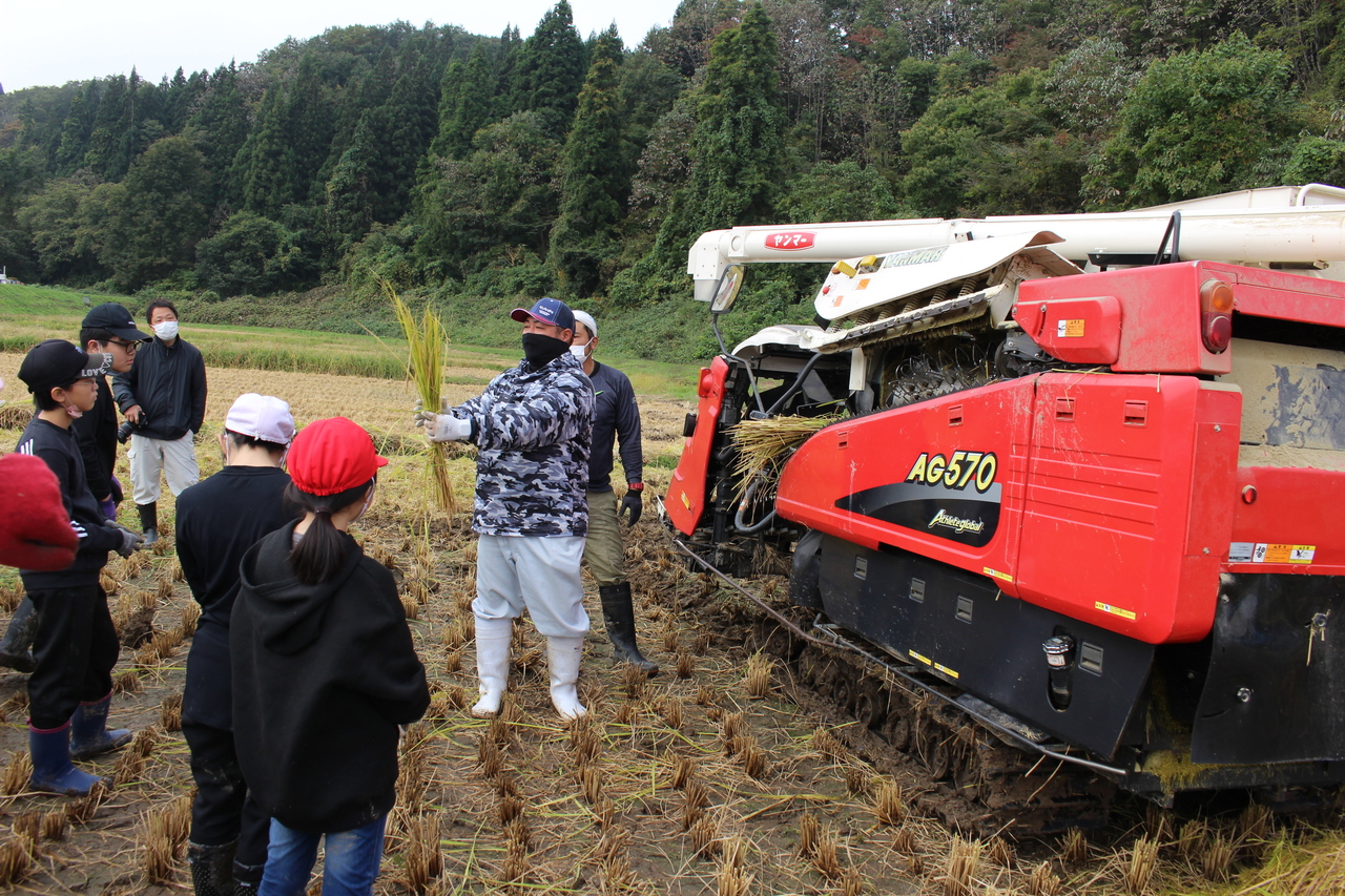 刈取機の横で長靴を履いた男性が、集まった子供たちに刈り取った稲を見せている写真
