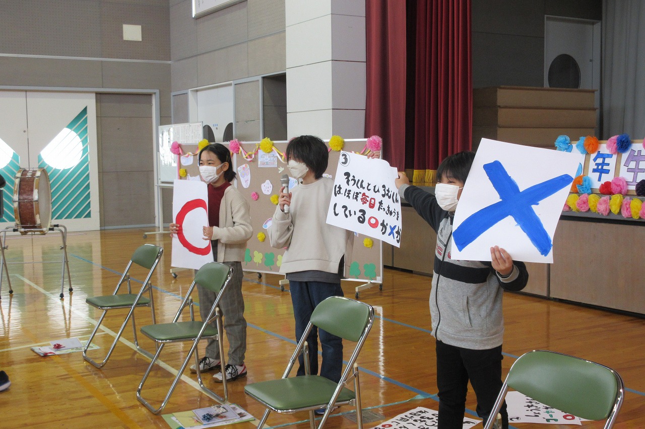 小学校の体育館にて行われた6年生を送る会にて、男子児童2人と女子児童1人が壇上の前で、まるとばつ、クイズの内容が書かれたフリップを持って、クイズの司会をしている様子を写した写真