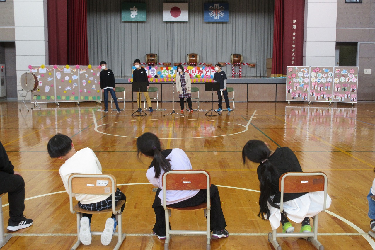 小学校の体育館にて行われた6年生を送る会にて、壇上の前で身体を揺らしながら歌を歌っている6年生の児童達に合わせながら、身体を揺らしている3人の児童の様子を写した写真