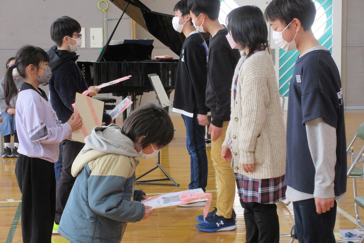 小学校の体育館にて行われた6年生を送る会にて、6年生の男子児童3人と女子児童1人に記念品を贈る児童達の様子を写した写真