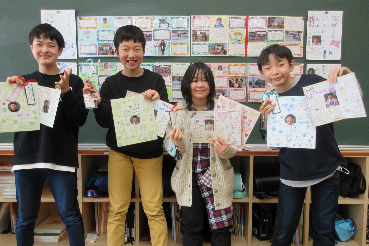 小学校の教室内にて、6年生を送る会で贈呈された記念品を見せながら、並んで笑顔を向ける6年生の男子児童3人と女子児童1人を写した記念写真