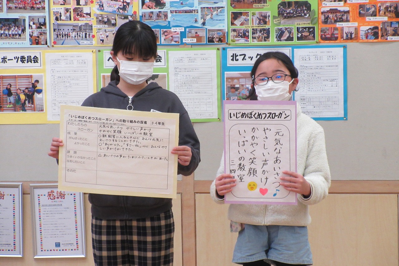 3・4年生のいじめぼくめつスローガンと取り組みの反省のプリントをみんなに見せるように広げて持っている2人の女子児童の写真