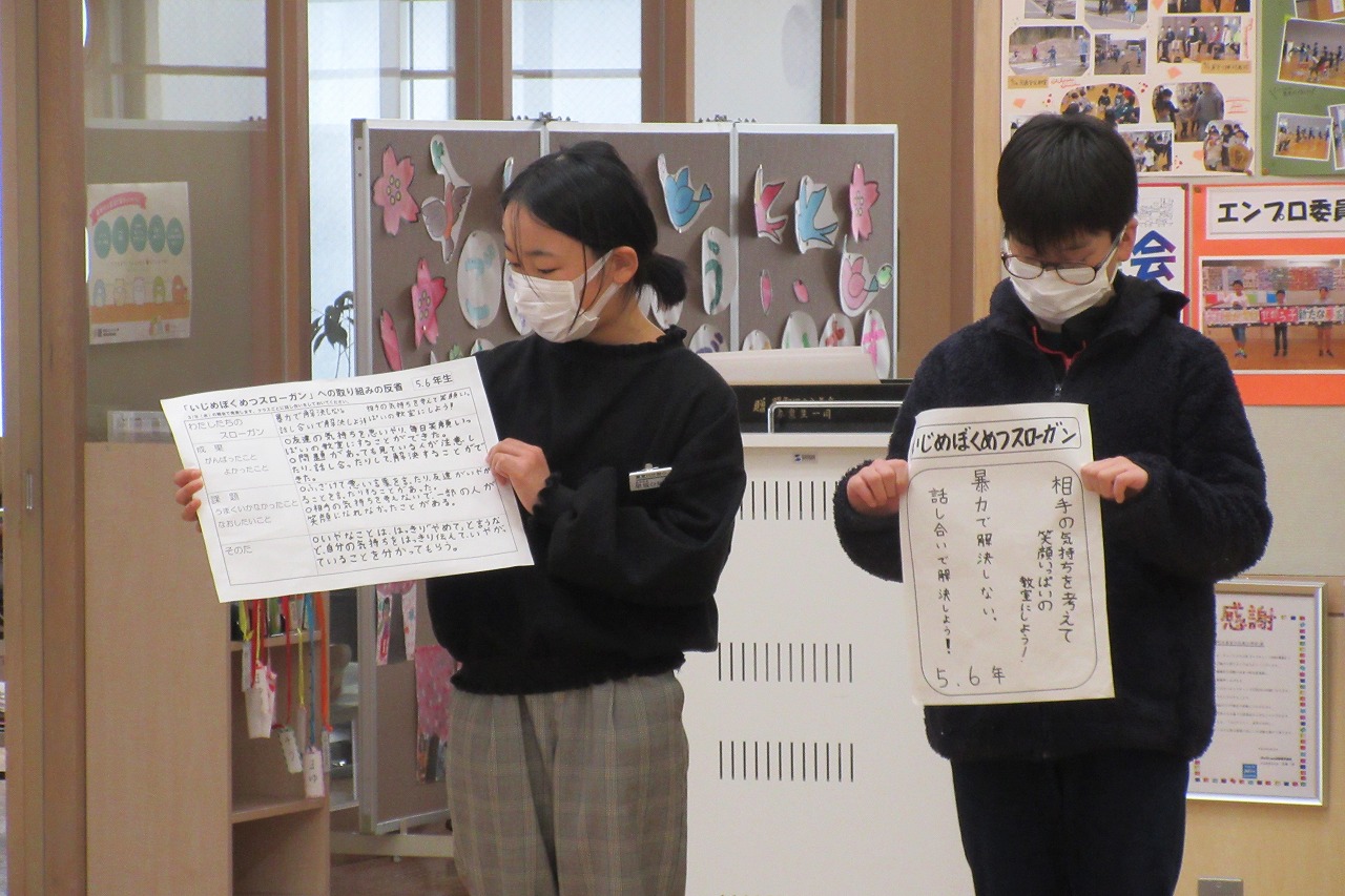 5・6年生のいじめぼくめつスローガンと取り組みの反省のプリントをみんなに見せながら発表している女子児童と男子児童の写真