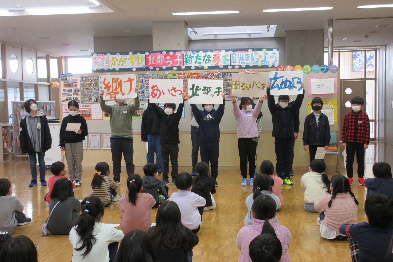 来年度のスローガン「響け あいさつ 北部っ子 明るい心を 広めよう」が5枚の紙に分けて書かれ、それを高々と掲げる児童たちの写真
