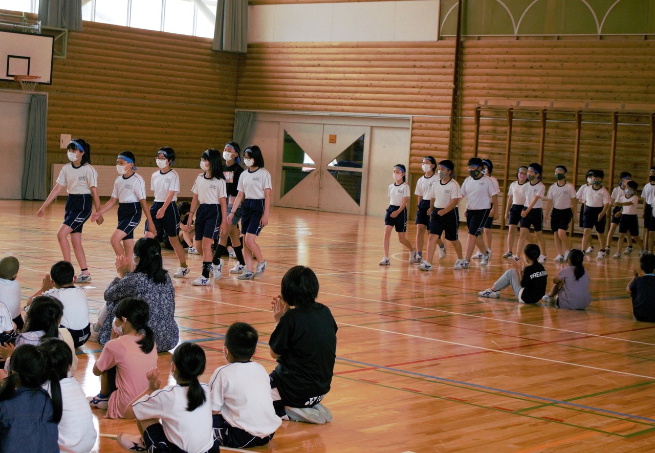 青い鉢巻を巻いて行進する子供たちと、床に座ってそれを眺める子供たちと大人の写真