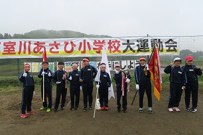 「真室川あさひ小学校大運動会」と書かれた横断幕の前で優勝旗と紅白旗を持つ児童たちの写真