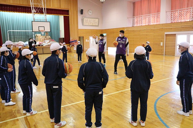 体育館で、プロバスケットボール選手のドリブルに関する説明を熱心に聴く児童たちの写真
