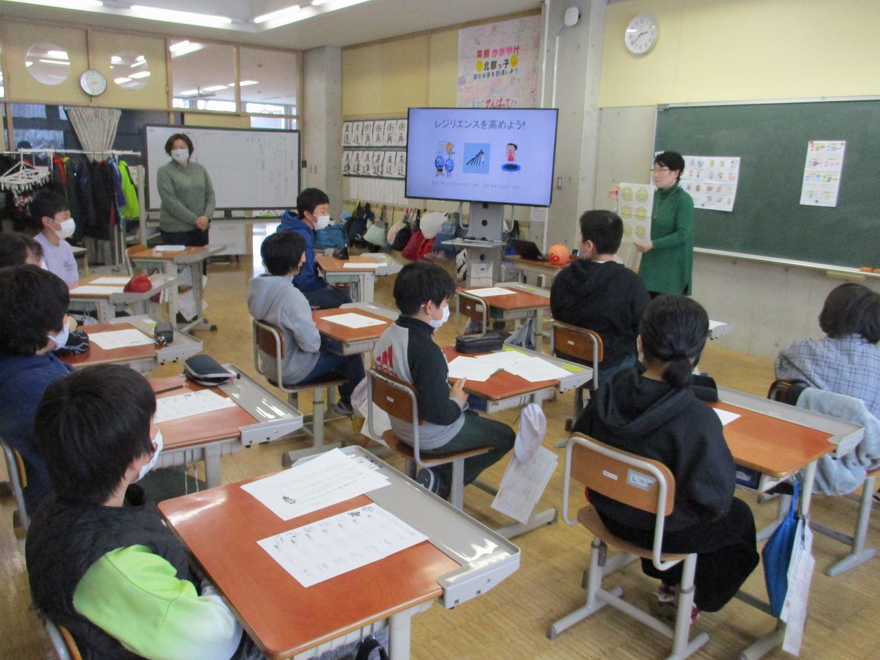レジリエンスについてモニターを使って説明するスクールカウンセラーとその話を椅子に座って聞く生徒たちの写真
