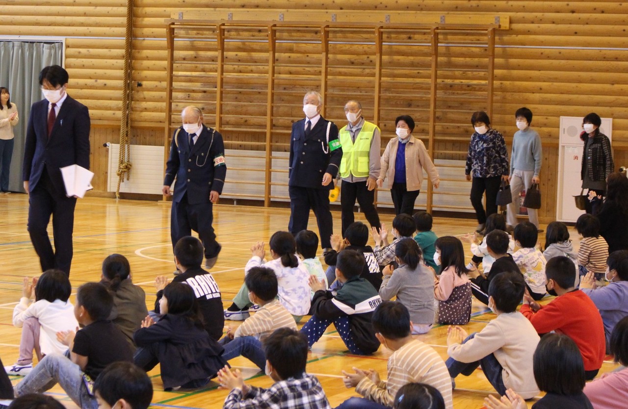 体育館で座った子供たちが入場する見守り隊の大人を迎え入れている写真
