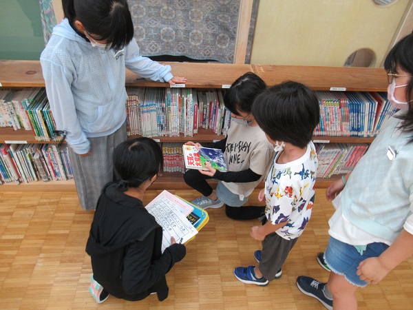 本棚の手前で子供たち5人が本を手に取ったりその表紙を一緒に見ている写真