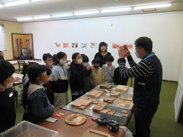 小さな木材が並んだ机の横で男性が話し、大勢の子供たちがその話を聞く写真