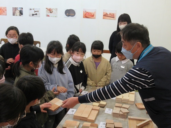 小さな木材が並んだ机の横に子供たちが集まり、その中央で木材を手に持つ子供と、手を伸ばす男性の写真