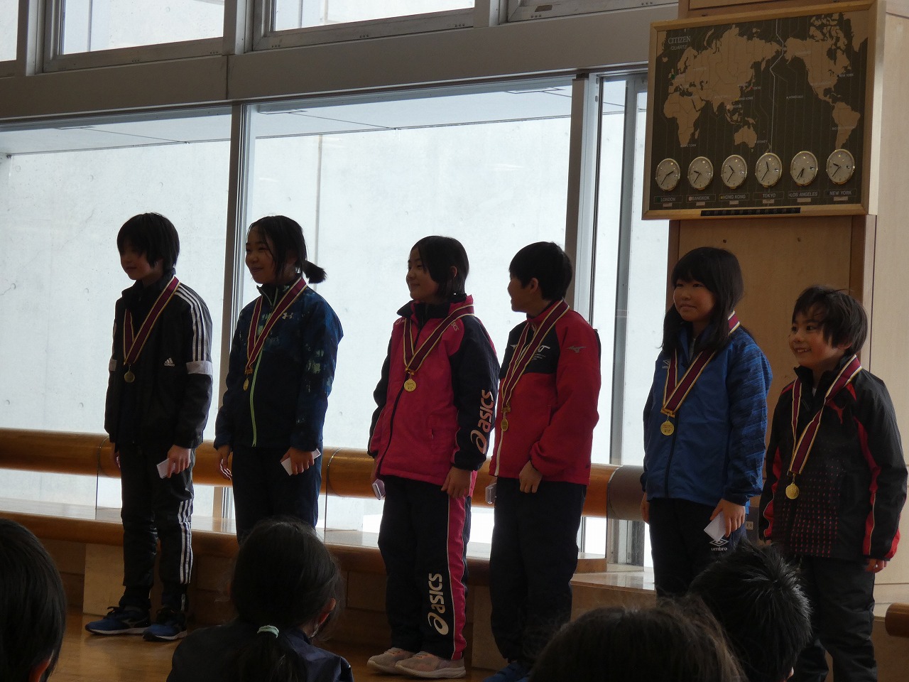 真室川北部小学校スキー大会にて、スキー場施設内で床に座る児童達に、向かい合ってメダルをかけて立っている、スキー大会参加者の児童6人の様子を写した写真