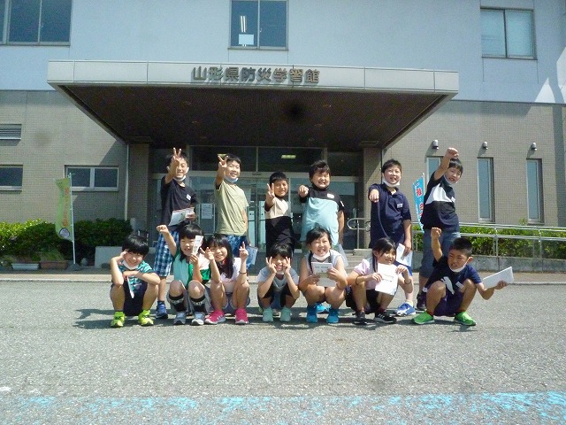校外学習にて、山形県防災学習館入口前で、2列に並んで各々ポーズを取っているところを撮影した、3年生の児童13人の記念写真