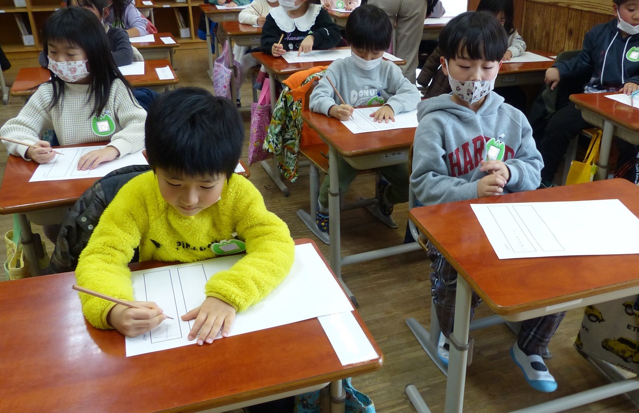 机に座って紙に文字を書こうとしている子供たちの写真