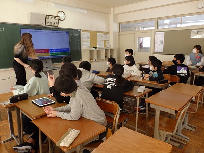 教室で席に着き、英語ネイティブスピーカーの女性から、モニター画面を使った講義を受ける児童たちの写真