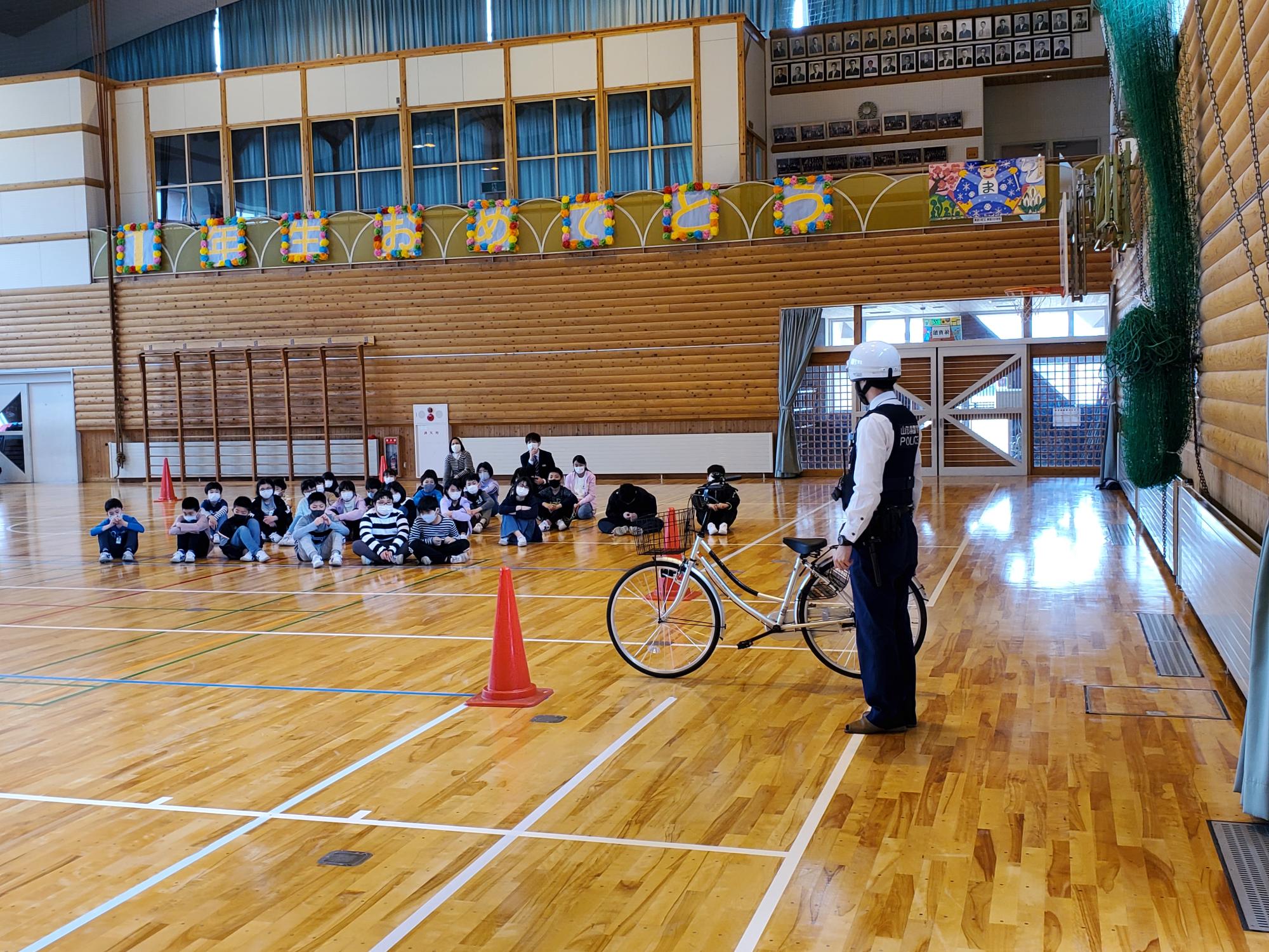 体育館で床に座る子供たちが自転車の横に立つ警察官に注目している写真