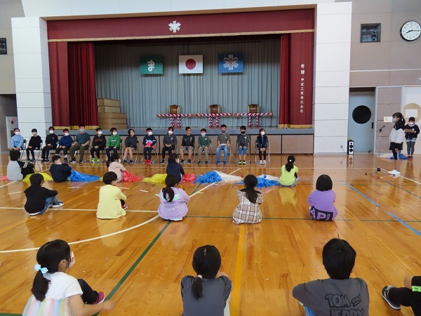 1列に並んだ椅子に腰掛ける子供たちと、体育座りをした子供たちがマイクに向かっている女の子を眺めている写真