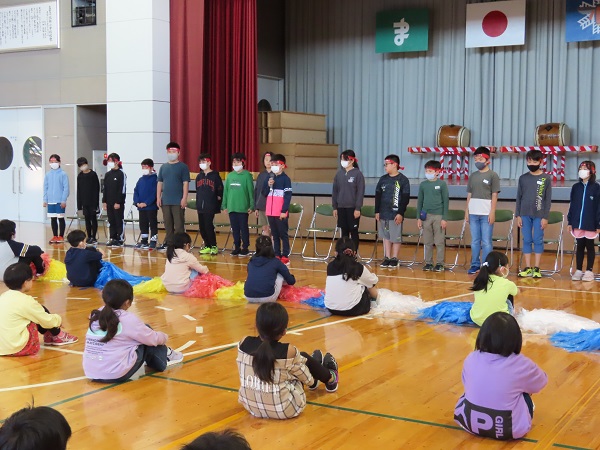 1列に並んだ椅子の前に整列する赤い鉢巻を巻いた子供たちと、その向かいで体育座りをする子供たちの写真