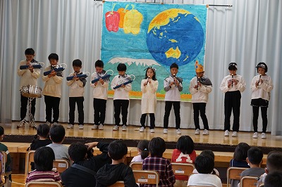 地球が描かれた垂れ幕の前で鍵盤ハーモニカやリコーダーで題名のない音楽かい？を演奏する5年生児童たちの写真