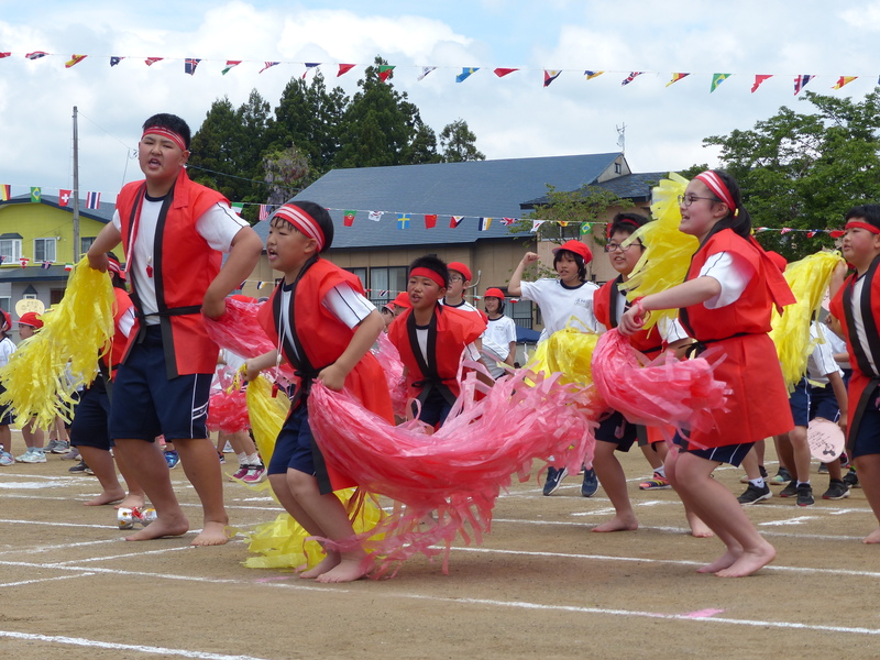 赤い法被を着て赤と黄色のポンポンを振りかざして踊る児童たちの写真
