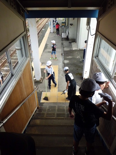 駅のホームを箒で掃く運動帽子を被った子供たちと、階段の手すりを拭く子供2人の写真