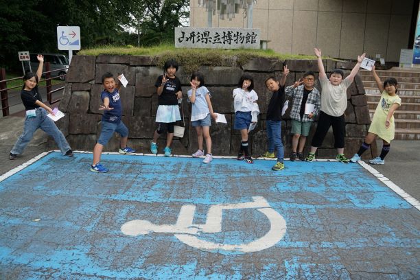 校外学習にて、山形県立博物館の入口前の駐車場で、4年生の児童9人が、各々ポーズを取っているところを撮影した記念写真