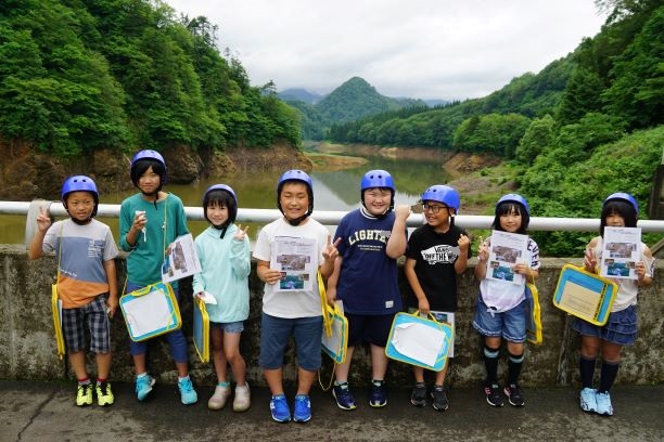 曇り空のもと、高坂ダムの天端で、周囲の緑豊かな自然を背景に、4年生の児童8人がピースサインをしているところを撮影した記念写真