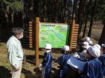 晴天の中村湿原の「中村湿原案内図」の前に立っているガイドの男性と、白い帽子に青いジャージ姿の3年生の児童達の様子を写した写真