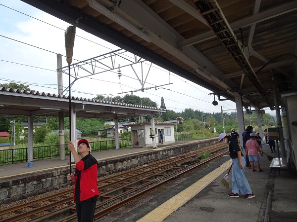 駅のホームで、長い箒を用いて天井を掃除する子供や、箒を持って地面を掃く子供たちの写真