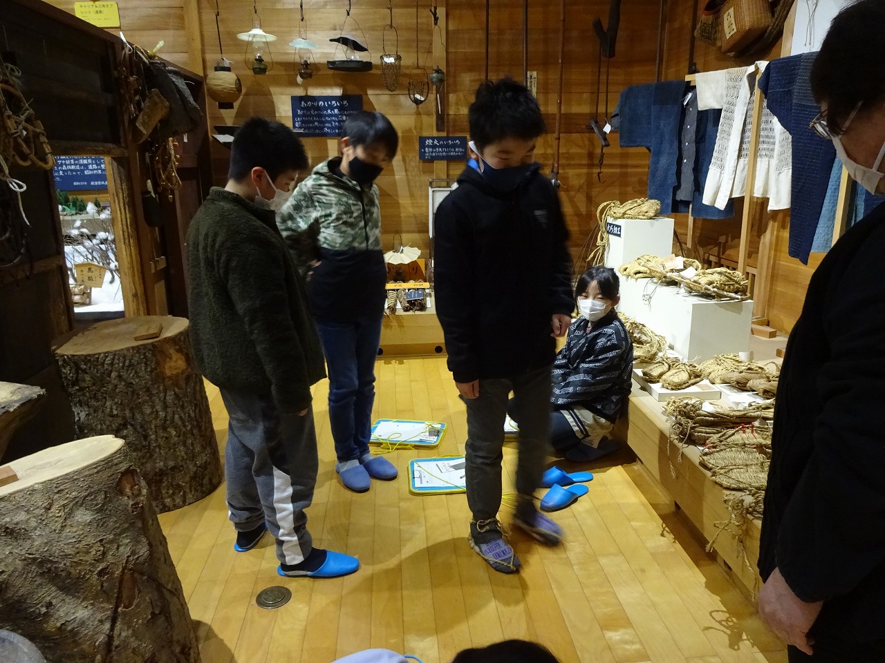 服や藁でできた靴が展示されている部屋で藁の靴の試し履きをしている生徒たちの写真