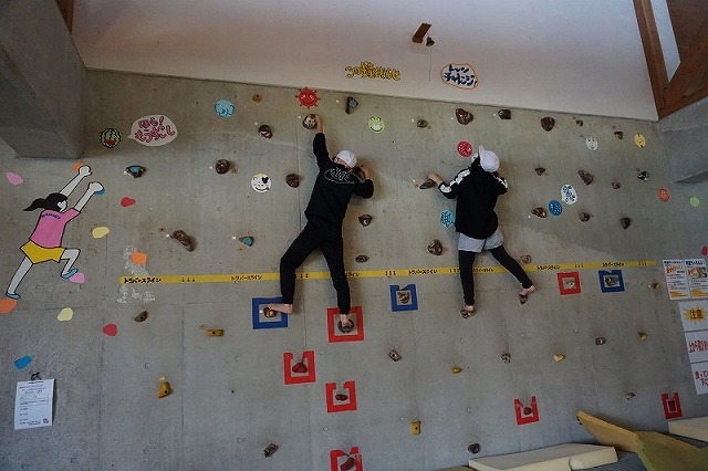 壁面の上部にある小さな鐘を先に鳴らすために、競い合ってクライミングをする二人の児童の写真