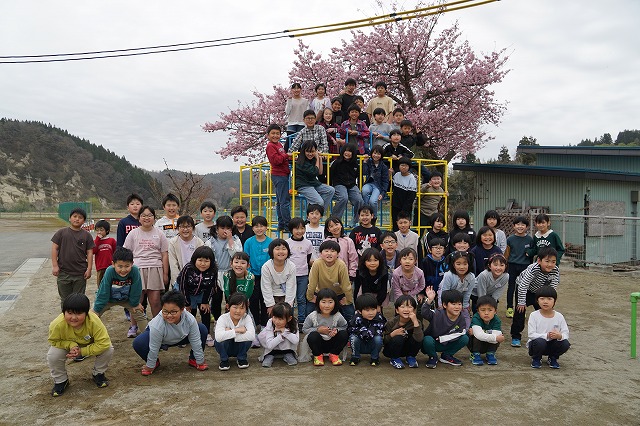 曇り空の小学校の校庭にて、後ろの方でジャングルジムに乗って笑顔を向けている児童達、その前の方で、4列に並んで笑顔を向けている大勢の児童達が写った集合写真