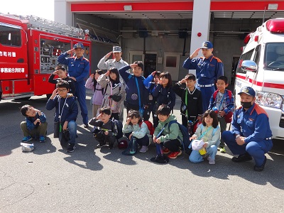 4人の消防ユニフォームを着た男性と子供たちが消防車の横で敬礼のポーズを取っている写真