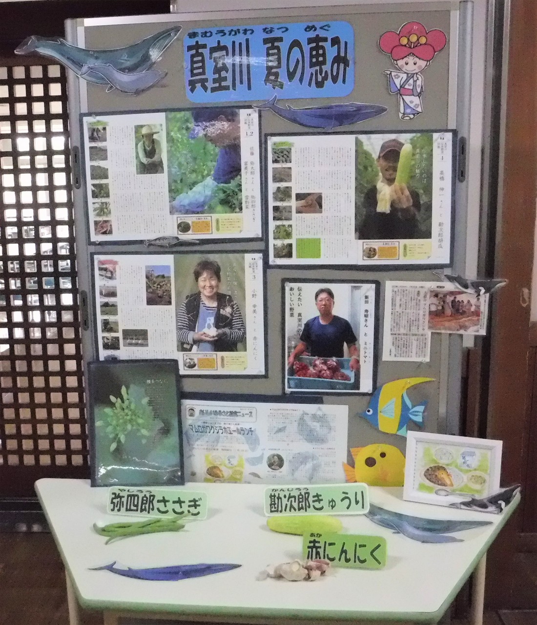「真室川夏の恵み」と書かれたパネルとその下に地元の野菜「弥四郎ささぎ」「勘次郎きゅうり」「赤にんにく」が展示されている写真