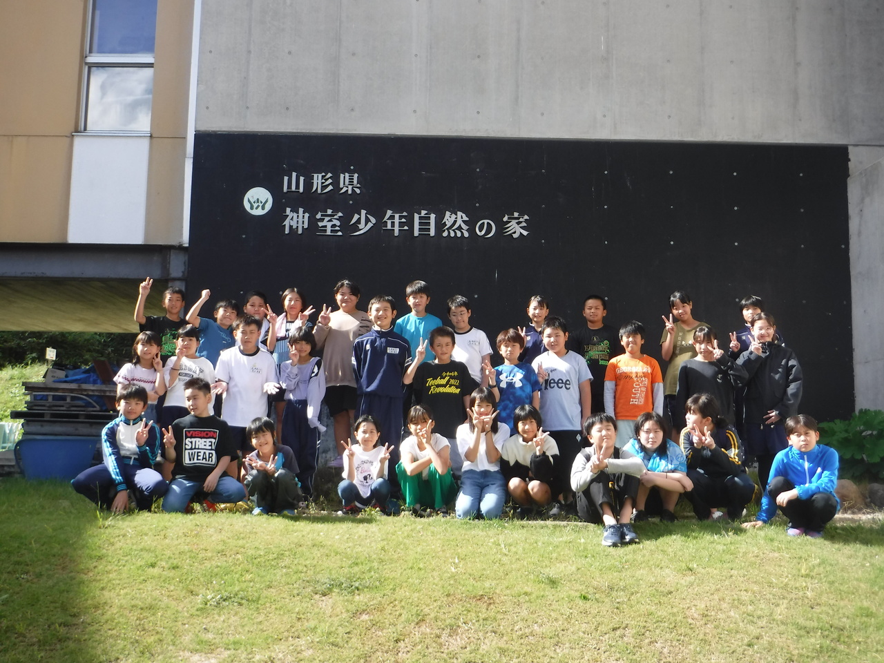 山形県神室少年自然の家と書かれた看板の前の集合しピースサインを向ける子供たちの写真