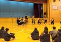 体育館の床に座った人達の前で楽器を持った複数の人が演奏をしている写真