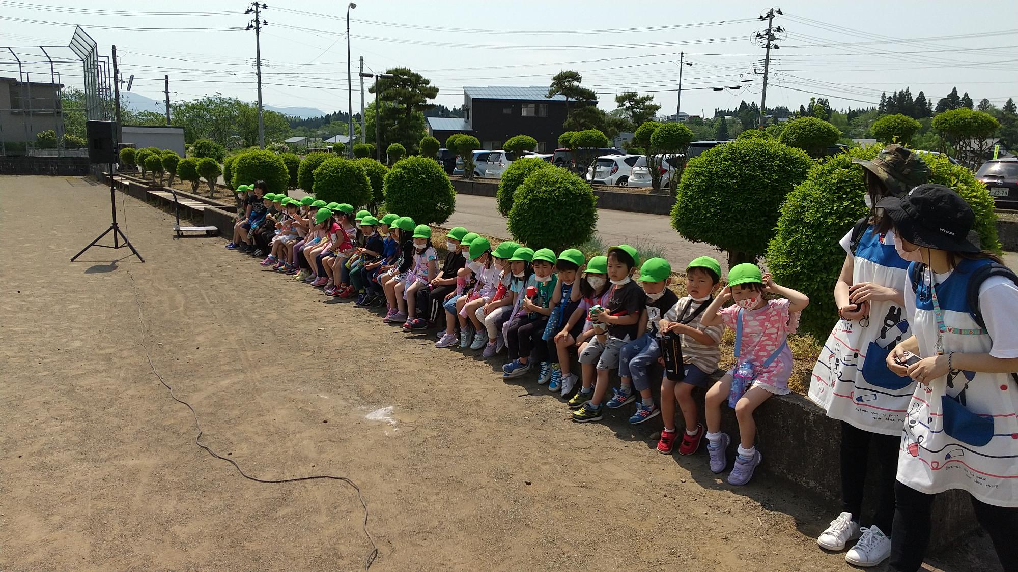 低い塀に1列に腰かける緑の運動帽を被った小さい子供たちと、列の端で子供と会話する帽子を被った女性2人の写真